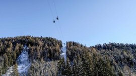 La Valle di Altafossa, presso Maranza L'immagine mostra una funivia con cabine che passano sopra alberi coperti di neve e una collina boscosa. Il cielo è sereno e blu, e la funivia conduce a una cima anch'essa coperta di neve.