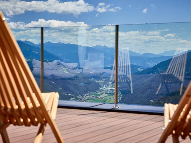 Benvenuti nell’Area Vacanze Sci e Malghe Rio Pusteria! L'immagine mostra due sdraio in legno su una terrazza con ringhiera in vetro, attraverso la quale si può vedere un vasto paesaggio montano. La vista mostra valli verdi, colline boscose e montagne blu sullo sfondo sotto un cielo leggermente nuvoloso.