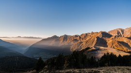 Quando si dice idilliaco… ecco a voi Fundres L'immagine mostra un ampio paesaggio montano all'alba o al tramonto. Le montagne sono immerse in una luce calda e dorata, mentre la nebbia riempie le valli. Sul bordo destro dell'immagine si vede un piccolo rifugio di montagna situato su una collina, anch'esso illuminato dalla luce soffusa.