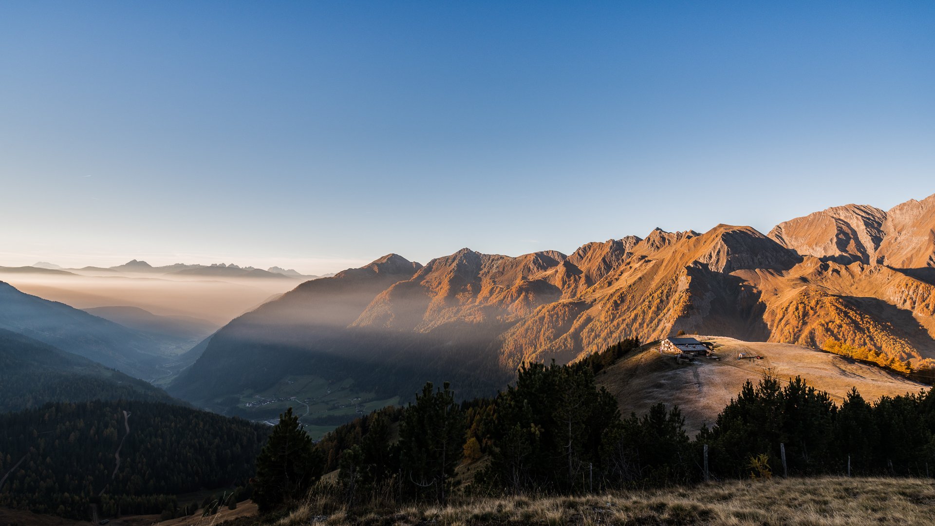 Quando si dice idilliaco… ecco a voi Fundres L'immagine mostra un ampio paesaggio montano all'alba o al tramonto. Le montagne sono immerse in una luce calda e dorata, mentre la nebbia riempie le valli. Sul bordo destro dell'immagine si vede un piccolo rifugio di montagna situato su una collina, anch'esso illuminato dalla luce soffusa.