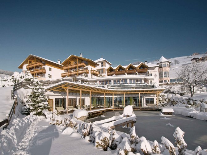 Benvenuti nell’Area Vacanze Sci e Malghe Rio Pusteria! L'immagine mostra un grande hotel in un paesaggio invernale coperto di neve. Davanti all'hotel c'è un laghetto ghiacciato, e i tetti così come l'ambiente circostante sono spessi di neve.