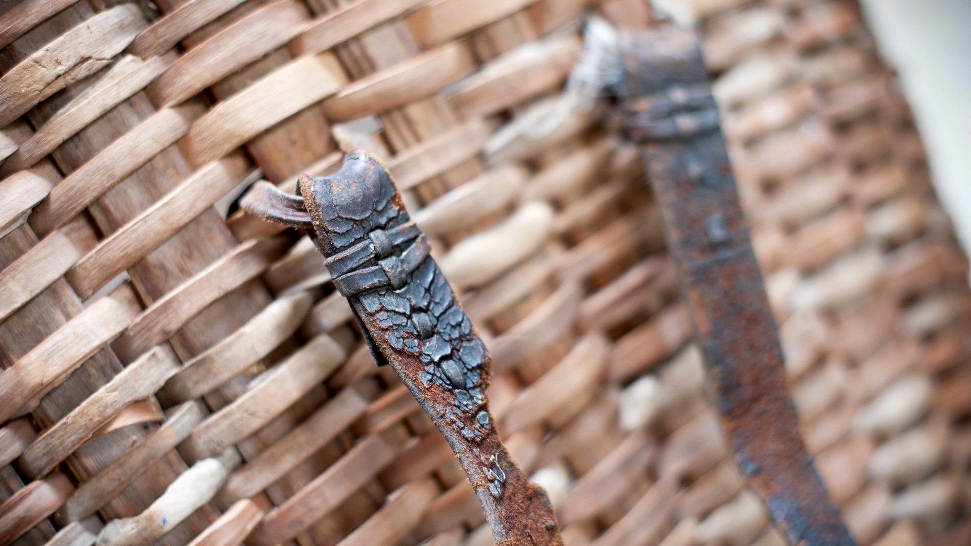 Il Sentiero dei masi di Fundres L'immagine mostra un primo piano di un cesto intrecciato fatto di strisce di legno. In primo piano si vedono due cinturini di metallo arrugginito fissati al cesto, con la struttura del cesto e le crepe nel metallo chiaramente visibili.