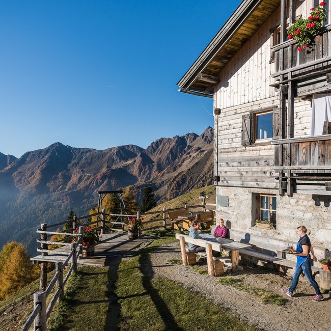 Gampiel Alm L'immagine mostra un tradizionale rifugio di montagna in legno, nel cui esterno gli ospiti godono della vista soleggiata sulle montagne autunnali.
