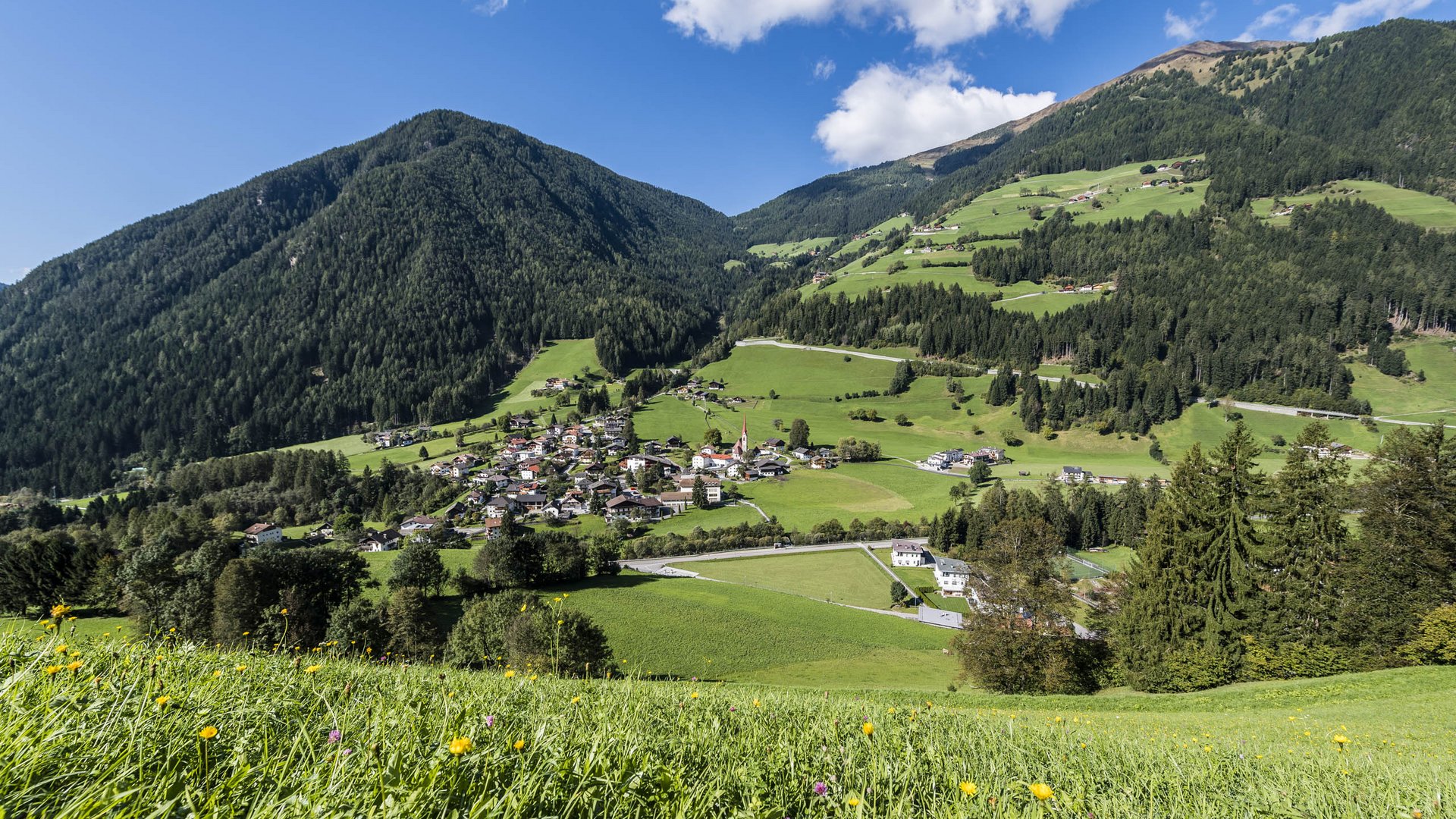 Quando si dice idilliaco… ecco a voi Fundres L'immagine mostra un paesaggio pittoresco con colline verdi e un piccolo villaggio incastonato in una valle. In primo piano ci sono prati fioriti, mentre lo sfondo è dominato da montagne boschive e un cielo blu con qualche nuvola.