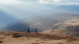 I percorsi più belli delle Dolomiti L'immagine mostra due escursionisti che attraversano una collina coperta d'erba. Sullo sfondo si estende un ampio paesaggio collinare sotto un cielo nuvoloso, attraverso il quale alcuni raggi di sole filtrano drammaticamente, immergendo la scena in una luce soffusa.
