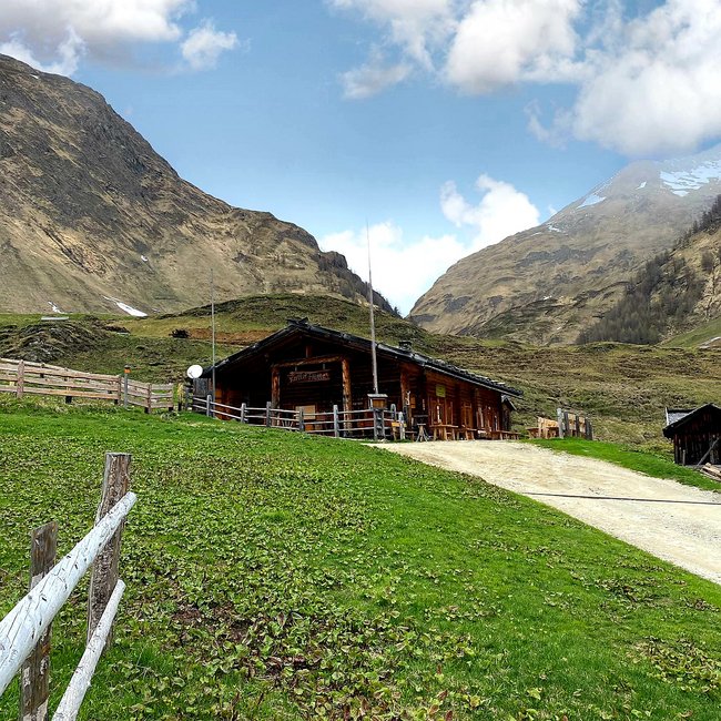 Kuttnhütte L'immagine mostra un rustico rifugio di montagna in legno, situato su una dolce collina verde. Il rifugio è circondato da una recinzione di legno e sullo sfondo si ergono alte montagne rocciose sotto un cielo azzurro con nuvole sparse.