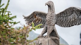 Il Parco del Sole Gitschberg – Maranza L'immagine mostra una scultura dettagliata in legno di un'aquila con le ali spiegate. In primo piano si vede della vegetazione, mentre lo sfondo è costituito da un cielo nuvoloso e montagne sfocate.