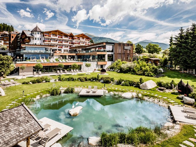 Benvenuti nell’Area Vacanze Sci e Malghe Rio Pusteria! L'immagine mostra un grande edificio alberghiero con più piani e balconi, circondato da un giardino ben curato. In primo piano c'è uno stagno naturale con lettini su piattaforme di legno, circondato da prato verde e pietre.