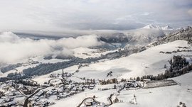 Vacanze sugli sci con i bambini a Terento L'immagine mostra un paesaggio innevato con un piccolo villaggio in primo piano e ampie colline sullo sfondo, ricoperte da boschi e campi. Sopra le colline si trovano nuvole dense, mentre sullo sfondo si vedono montagne innevate.