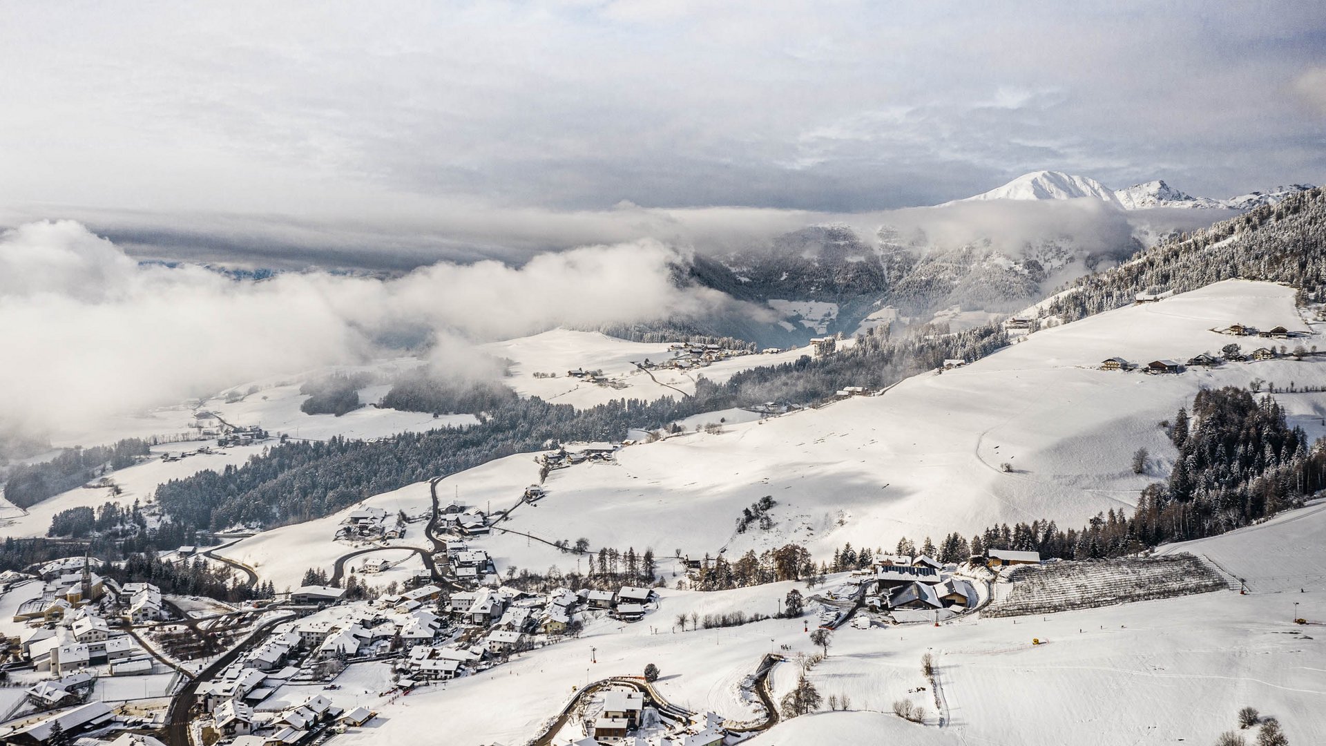Vacanze sugli sci con i bambini a Terento L'immagine mostra un paesaggio innevato con un piccolo villaggio in primo piano e ampie colline sullo sfondo, ricoperte da boschi e campi. Sopra le colline si trovano nuvole dense, mentre sullo sfondo si vedono montagne innevate.