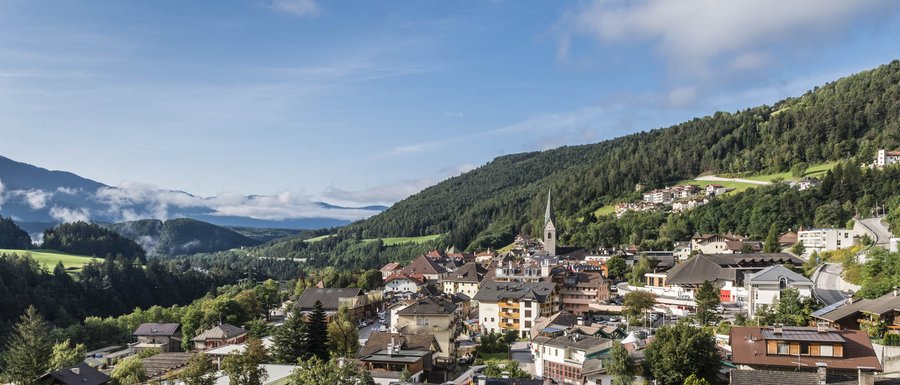 Le località dell’area vacanze Rio Pusteria L'immagine mostra una piccola città pittoresca in una valle verde, circondata da colline boscose e montagne. Al centro dell'immagine si erge un campanile con una cuspide appuntita, e gli edifici sono circondati da una vegetazione rigogliosa e alcune nuvole sparse.