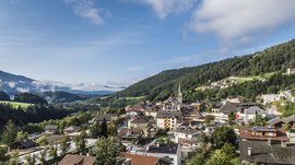 Alla scoperta di Rio di Pusteria L'immagine mostra una piccola città pittoresca in una valle verde, circondata da colline boscose e montagne. Al centro dell'immagine si erge un campanile con una cuspide appuntita, e gli edifici sono circondati da una vegetazione rigogliosa e alcune nuvole sparse.