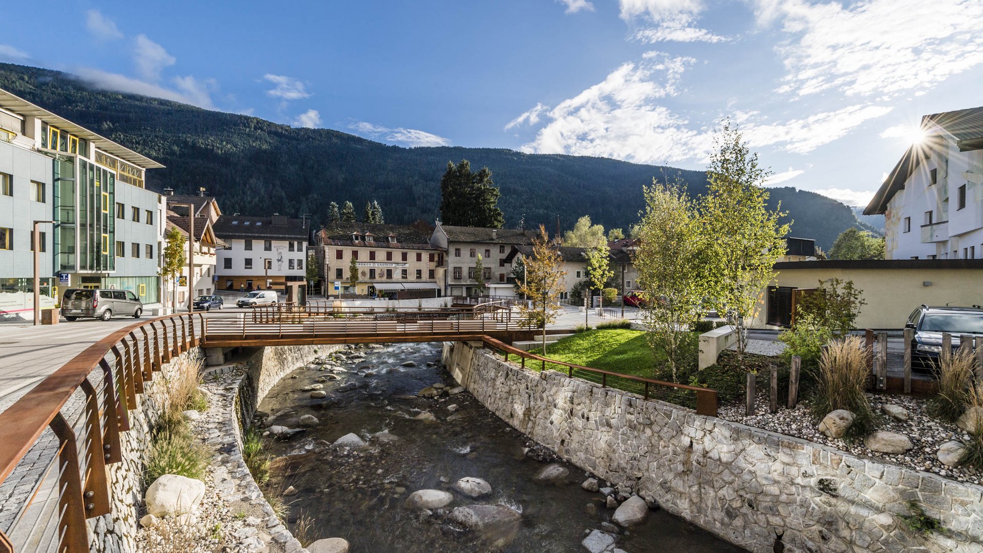 Vandoies: il cuore artigianale della Val Pusteria L'immagine mostra un piccolo ponte che attraversa un ruscello limpido in un villaggio. Sullo sfondo si vedono case e colline o montagne verdi sotto un cielo blu con poche nuvole.