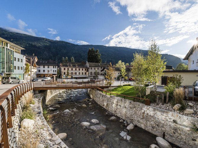 Le località dell’area vacanze Rio Pusteria L'immagine mostra un piccolo ponte che attraversa un ruscello limpido in un villaggio. Sullo sfondo si vedono case e colline o montagne verdi sotto un cielo blu con poche nuvole.