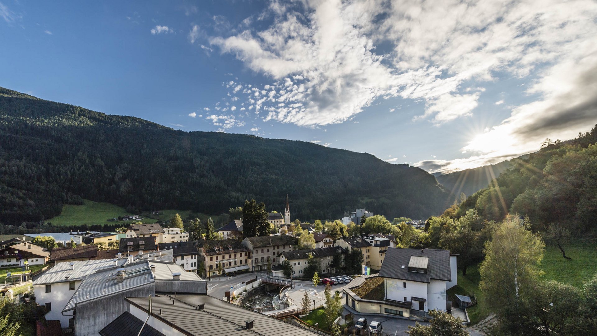Vandoies: il cuore artigianale della Val Pusteria L'immagine mostra una piccola città, incastonata tra colline boscose, con alcuni edifici in primo piano e una chiesa con una torre alta al centro. Il sole splende sopra le colline e proietta lunghe raggi di luce sul paesaggio, mentre il cielo è coperto da alcune nuvole.