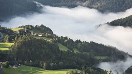 Il paradiso escursionistico di Rodengo L'immagine mostra un paesaggio collinare attraversato da una densa nebbia bianca. Su una delle colline si trova una piccola chiesa, circondata da prati verdi e boschi che emergono dalla nebbia.
