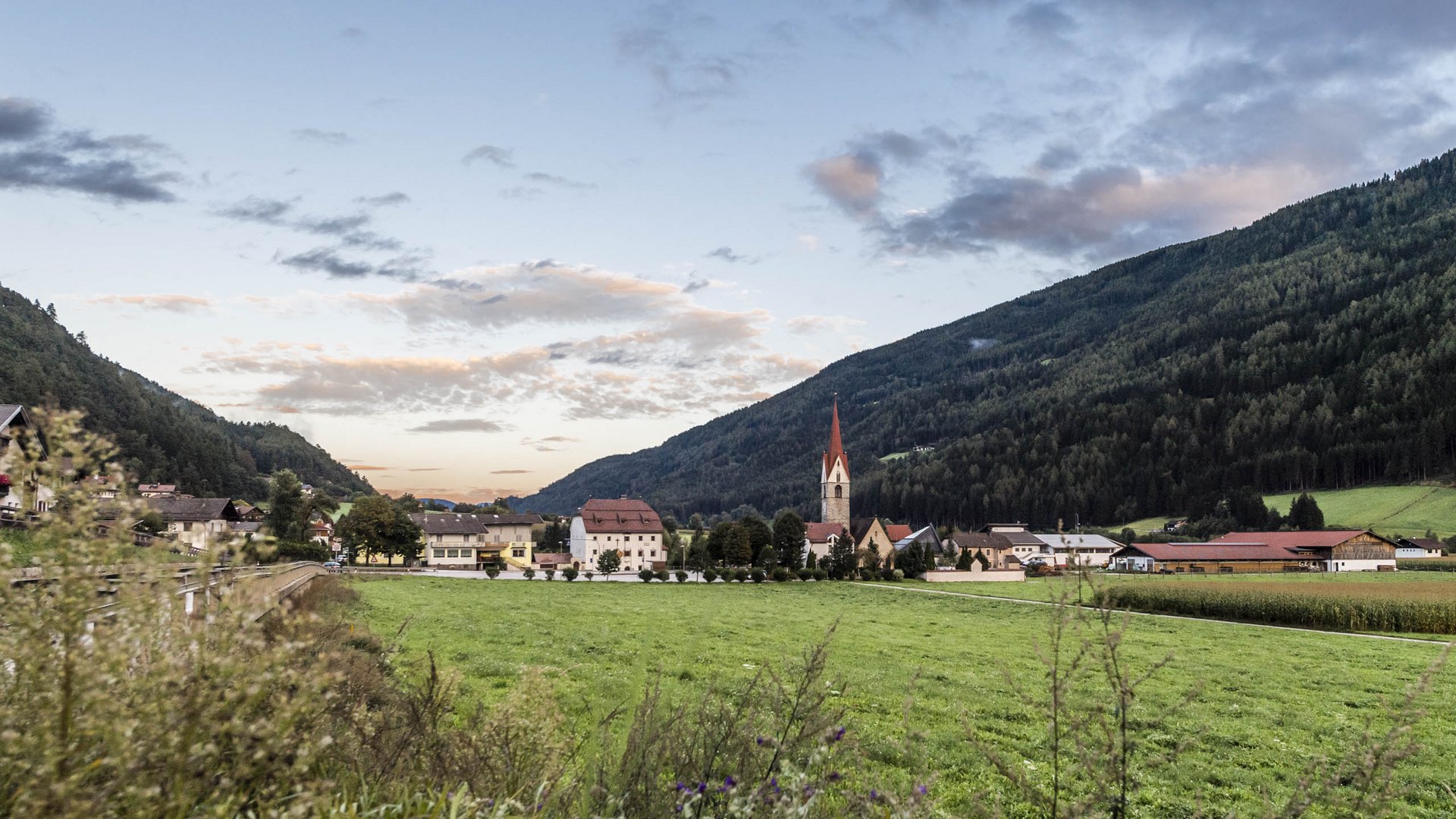 Vandoies: il cuore artigianale della Val Pusteria L'immagine mostra un'ampia prateria verde con un piccolo villaggio sullo sfondo, il cui centro è costituito da una chiesa con una torre alta e appuntita. Il villaggio è circondato da colline boschive sotto un cielo nuvoloso al tramonto.