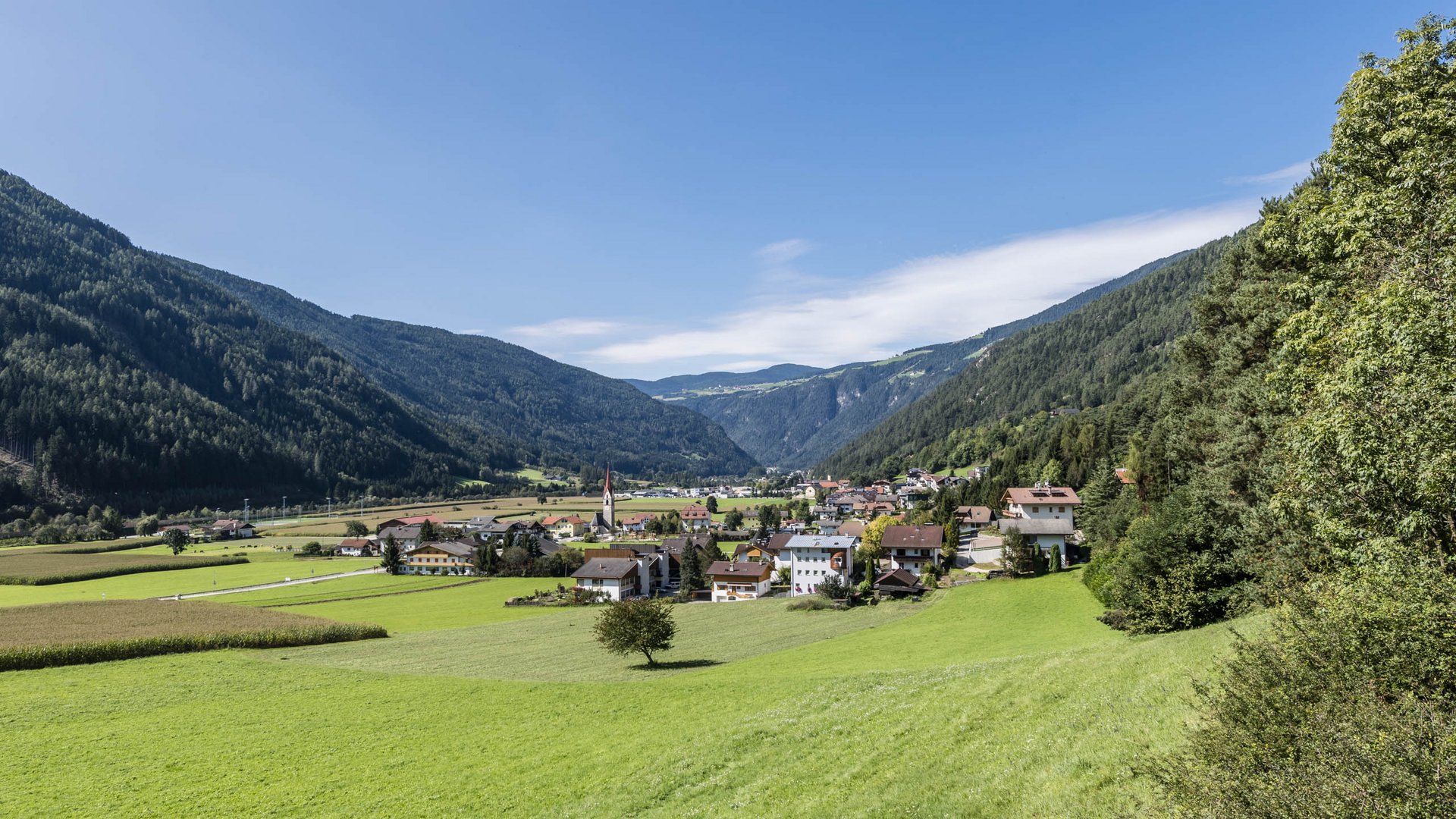 Vandoies: il cuore artigianale della Val Pusteria L'immagine mostra una valle verde con un piccolo villaggio in primo piano. Al centro del villaggio c'è una chiesa con una torre alta rossa, circondata da case residenziali e prati rigogliosi, mentre colline boschive circondano la valle.