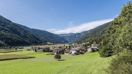 Vandoies: il cuore artigianale della Val Pusteria L'immagine mostra una valle verde con un piccolo villaggio in primo piano. Al centro del villaggio c'è una chiesa con una torre alta rossa, circondata da case residenziali e prati rigogliosi, mentre colline boschive circondano la valle.