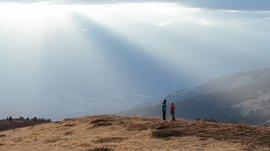 I percorsi più belli delle Dolomiti L'immagine mostra due escursionisti che stanno su una collina erbosa, circondati da un paesaggio montuoso vasto. Raggi di luce solare filtrano drammaticamente attraverso le nuvole e illuminano la nebbia in lontananza, creando una scena atmosferica e pacifica.
