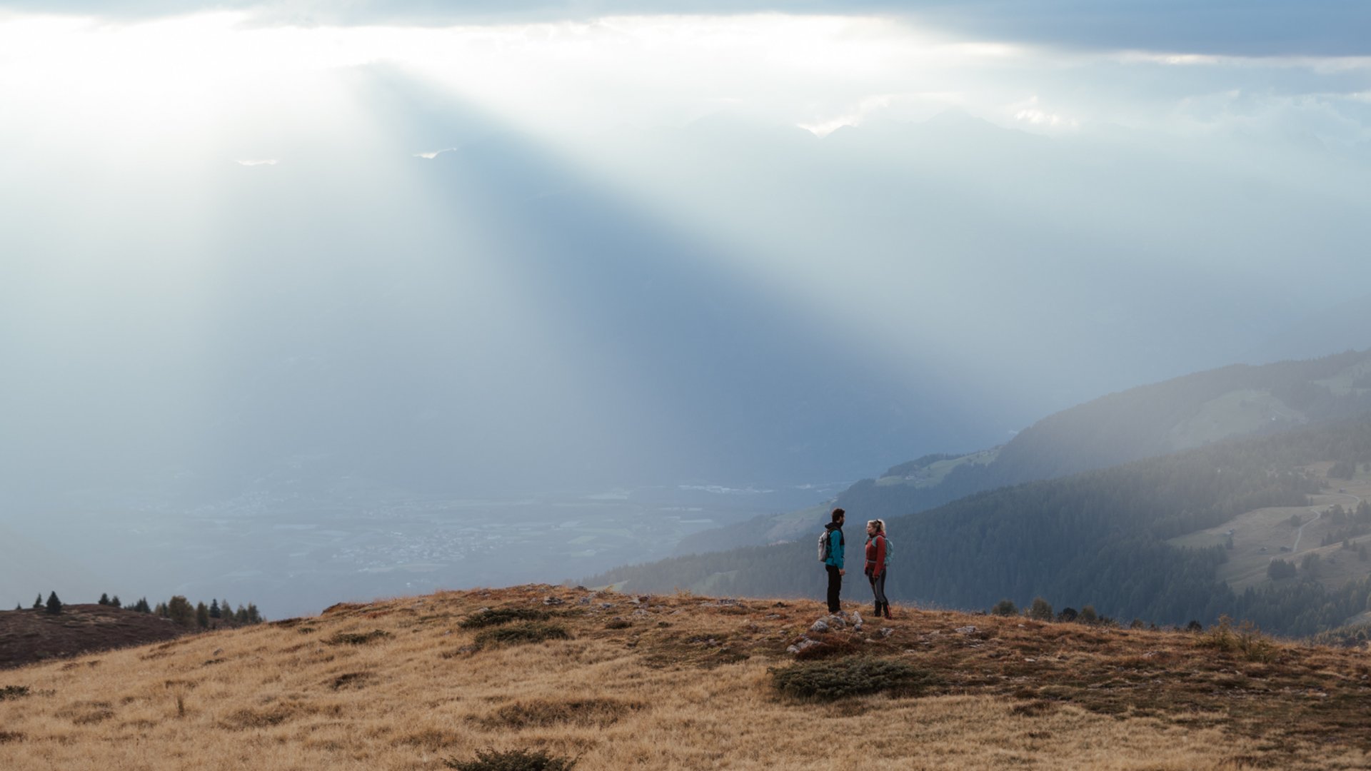 I percorsi più belli delle Dolomiti L'immagine mostra due escursionisti che stanno su una collina erbosa, circondati da un paesaggio montuoso vasto. Raggi di luce solare filtrano drammaticamente attraverso le nuvole e illuminano la nebbia in lontananza, creando una scena atmosferica e pacifica.