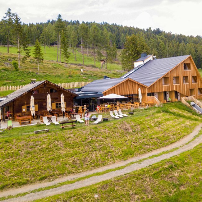 Oberhauserhütte L'immagine mostra un moderno rifugio di montagna in legno, circondato da prati verdi e foreste. Davanti al rifugio ci sono numerose sedie a sdraio e tavoli con ombrelloni, che invitano a rilassarsi nella natura.