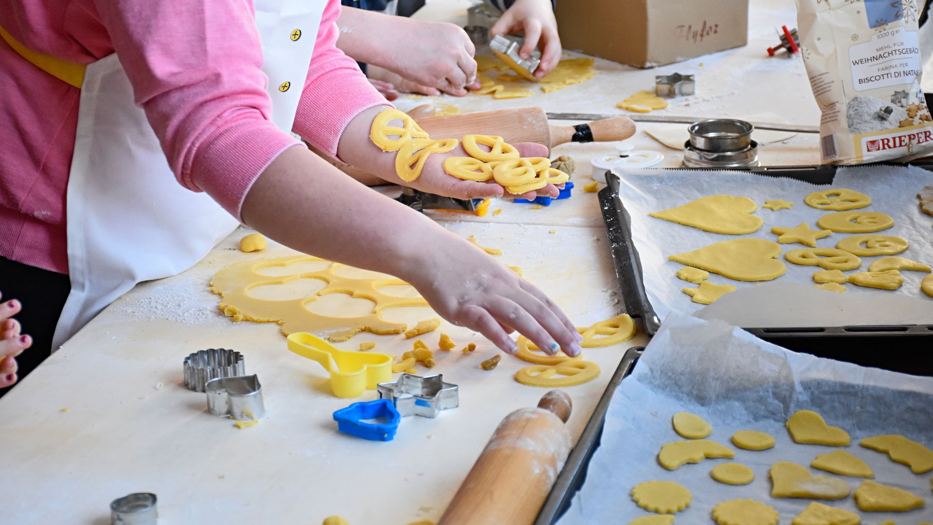 L’Avvento di Terento L'immagine mostra persone che fanno biscotti con delle formine per biscotti dall'impasto. Su un tavolo ci sono avanzi di impasto, formine per biscotti e un mattarello, mentre i biscotti ritagliati sono disposti su una teglia da forno in preparazione.
