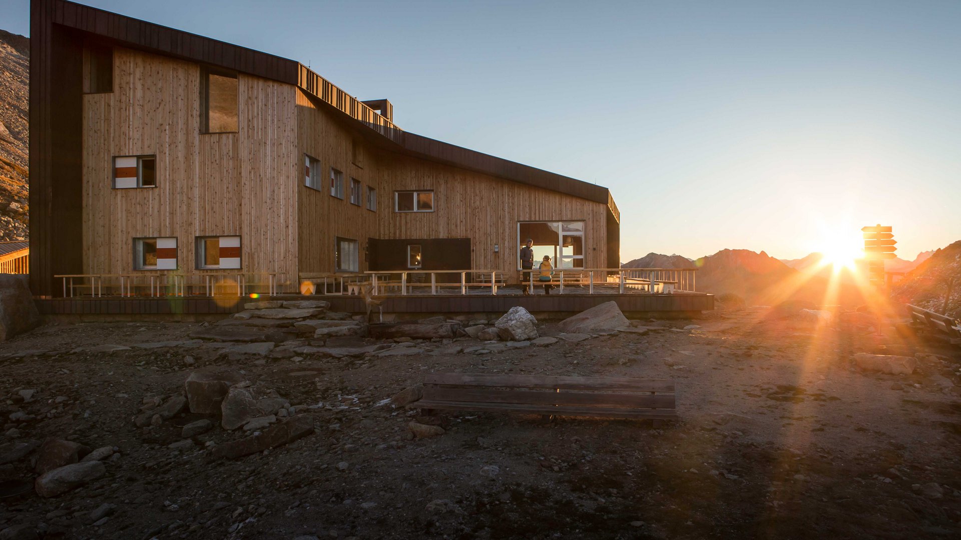 L’Alta via di Fundres L'immagine mostra un moderno rifugio di montagna in legno tra le montagne al tramonto. Il rifugio ha grandi finestre e una terrazza, mentre il sole tramonta dietro le montagne e avvolge l'ambiente in una luce calda.