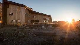 L’Alta via di Fundres L'immagine mostra un moderno rifugio di montagna in legno tra le montagne al tramonto. Il rifugio ha grandi finestre e una terrazza, mentre il sole tramonta dietro le montagne e avvolge l'ambiente in una luce calda.