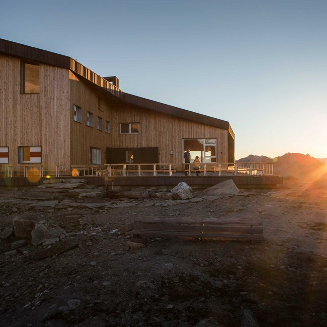 Edelrauthütte L'immagine mostra un moderno rifugio di montagna in legno tra le montagne al tramonto. Il rifugio ha grandi finestre e una terrazza, mentre il sole tramonta dietro le montagne e avvolge l'ambiente in una luce calda.
