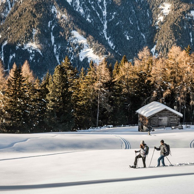 Rio Pusteria: escursioni invernali a Maranza e dintorni Due persone camminano nella neve davanti a una baita e alberi di conifere