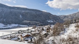 Vandoies: il cuore artigianale della Val Pusteria L'immagine mostra un villaggio innevato situato in una valle tra montagne densamente boscose e coperte di neve. In primo piano si vedono alcune case con il tetto coperto di neve, e sullo sfondo si erge una grande montagna boscosa sotto un cielo azzurro con poche nuvole.