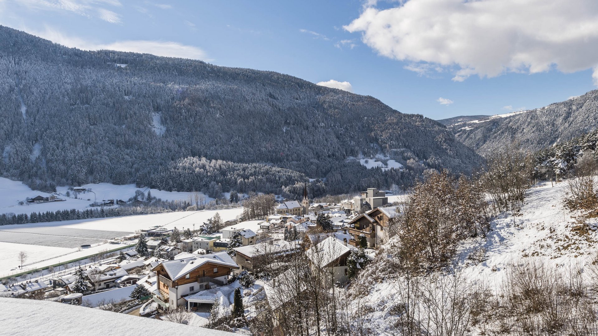 Vandoies: il cuore artigianale della Val Pusteria L'immagine mostra un villaggio innevato situato in una valle tra montagne densamente boscose e coperte di neve. In primo piano si vedono alcune case con il tetto coperto di neve, e sullo sfondo si erge una grande montagna boscosa sotto un cielo azzurro con poche nuvole.