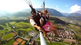 Parapendio in Alto Adige L'immagine mostra due persone che volano insieme in parapendio nell'aria. Sotto di loro si estende un paesaggio verde con campi, un campo sportivo e una piccola città. Le persone sembrano divertirsi, e il pilota tiene una fotocamera o un selfie stick per catturare il momento.