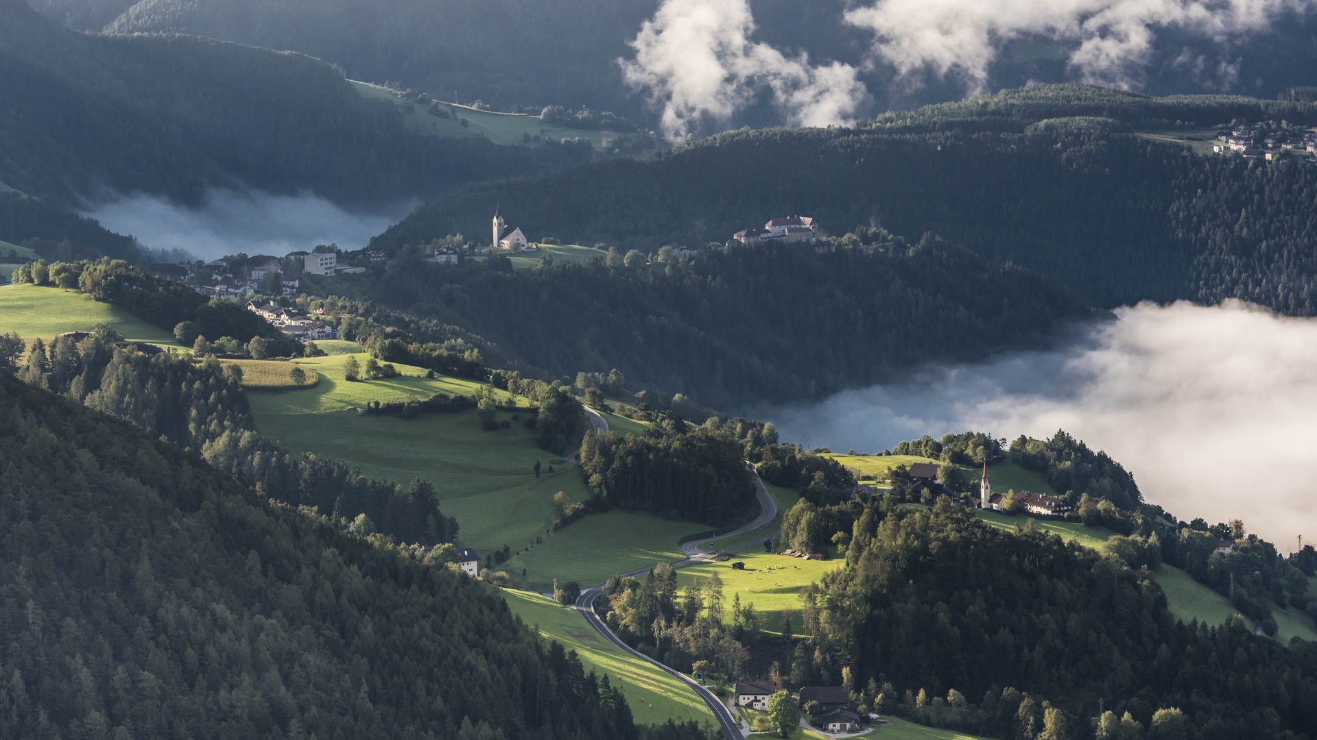 Il paradiso escursionistico di Rodengo L'immagine mostra un paesaggio collinare con prati verdi e foreste, attraversato da una strada tortuosa. Sullo sfondo si vedono una chiesa e un piccolo villaggio, circondati dalla nebbia che si insinua nelle valli.