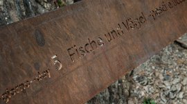 Il Sentiero della Creazione a Rodengo L'immagine mostra una lastra di metallo arrugginita con inciso il testo "Tag 5 pesci e uccelli". Lo sfondo è composto da elementi naturali come terra, pietre e corteccia d'albero.