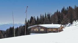 Escursioni a Spinga: qualche consiglio L'immagine mostra la "Anratter Hütte", una baita di legno su un pendio innevato, circondata da conifere. Sullo sfondo si vedono cime innevate sotto un cielo azzurro. Una bandiera rossa sventola su uno dei pennoni davanti alla baita.