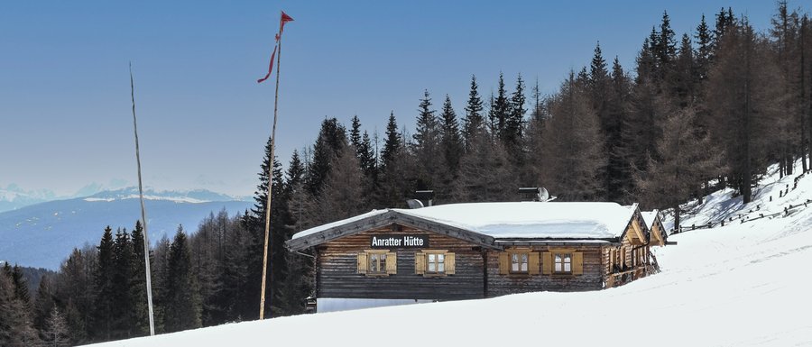 Le località dell’area vacanze Rio Pusteria L'immagine mostra la "Anratter Hütte", una baita di legno su un pendio innevato, circondata da conifere. Sullo sfondo si vedono cime innevate sotto un cielo azzurro. Una bandiera rossa sventola su uno dei pennoni davanti alla baita.