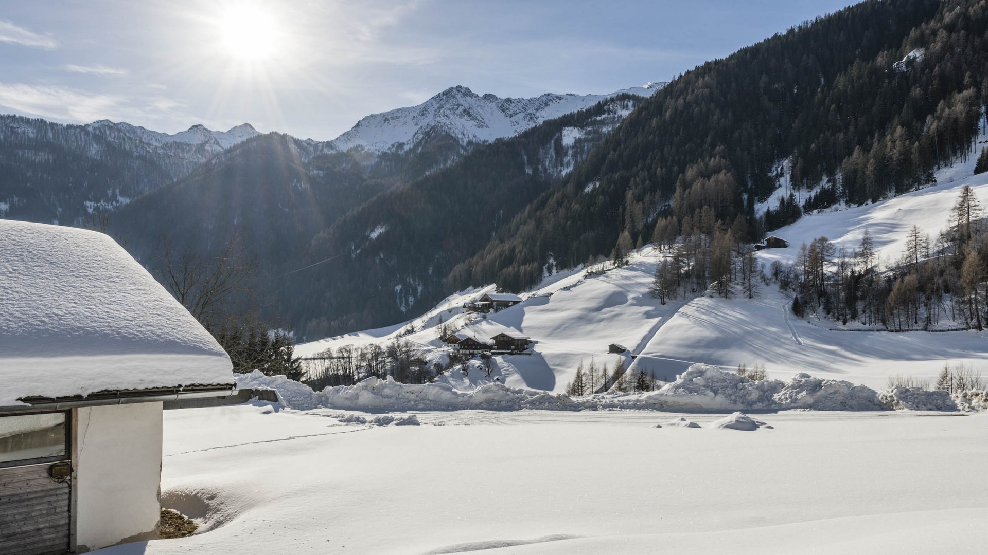 Quando si dice idilliaco… ecco a voi Fundres L'immagine mostra un paesaggio innevato con colline e montagne sullo sfondo. In primo piano si vede un edificio con un tetto coperto di neve, mentre il sole splende luminoso nel cielo irradiando le colline innevate.