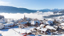 Vacanze sugli sci con i bambini a Terento L'immagine mostra un villaggio innevato con case e una chiesa in mezzo a un paesaggio montano invernale. Sullo sfondo, delle nebbie attraversano la valle, mentre il cielo è limpido e azzurro.