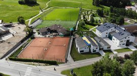 Il Parco Active a Terento L'immagine mostra una vista aerea di impianti sportivi in un ambiente rurale. Sono visibili due campi da tennis, un grande campo da calcio e alcuni edifici, tra cui scuderie con cavalli sul lato sinistro. L'ambiente è composto da prati verdi e colline.