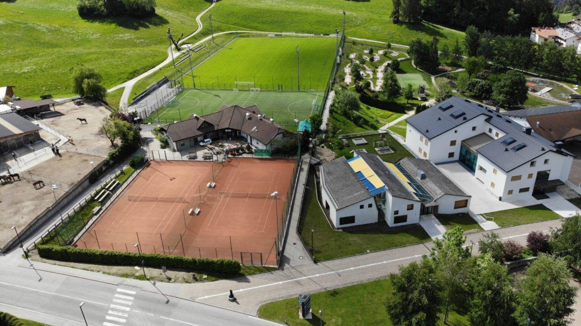 Il Parco Active a Terento L'immagine mostra una vista aerea di impianti sportivi in un ambiente rurale. Sono visibili due campi da tennis, un grande campo da calcio e alcuni edifici, tra cui scuderie con cavalli sul lato sinistro. L'ambiente è composto da prati verdi e colline.