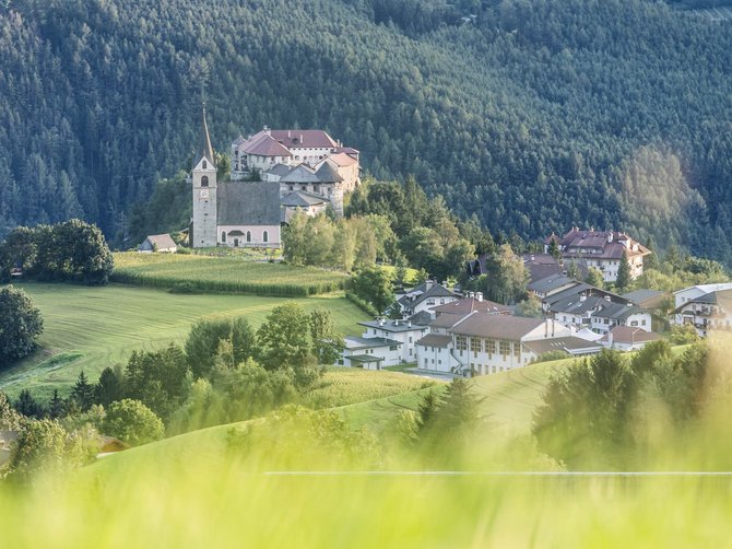 Le località dell’area vacanze Rio Pusteria L'immagine mostra una chiesa e un edificio su una collina, circondati da prati verdi e foreste dense. In primo piano si vede dell'erba sfocata.