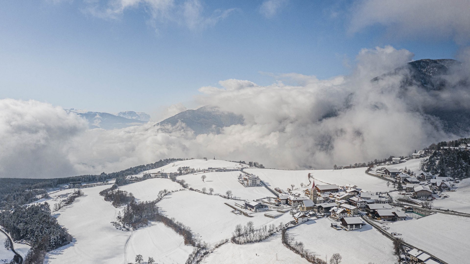 La frazione montana di Spinga L'immagine mostra un paesaggio montano innevato con un piccolo villaggio in primo piano. Il cielo è limpido e nuvole si muovono sulle montagne sullo sfondo, mentre le case e i campi del villaggio sono coperti di neve.