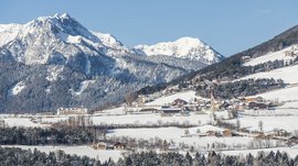 La frazione montana di Spinga L'immagine mostra un paesaggio invernale con un villaggio innevato in primo piano e montagne coperte di neve sullo sfondo. Il villaggio è circondato da boschi e prati, e una chiesa con una torre rossa spicca.
