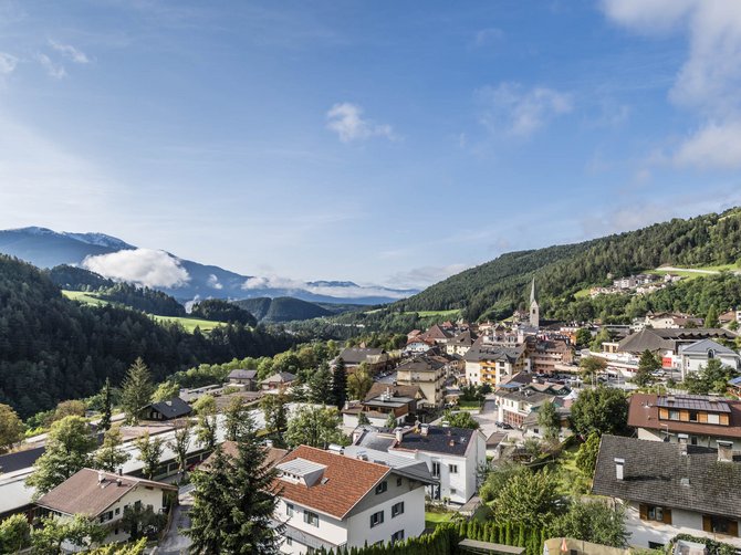 Le località dell’area vacanze Rio Pusteria L'immagine mostra una pittoresca città circondata da verdi colline e montagne. In primo piano si vedono case con tetti rossi e vegetazione rigogliosa, mentre sullo sfondo si riconoscono montagne avvolte dalla nebbia e un cielo azzurro limpido.