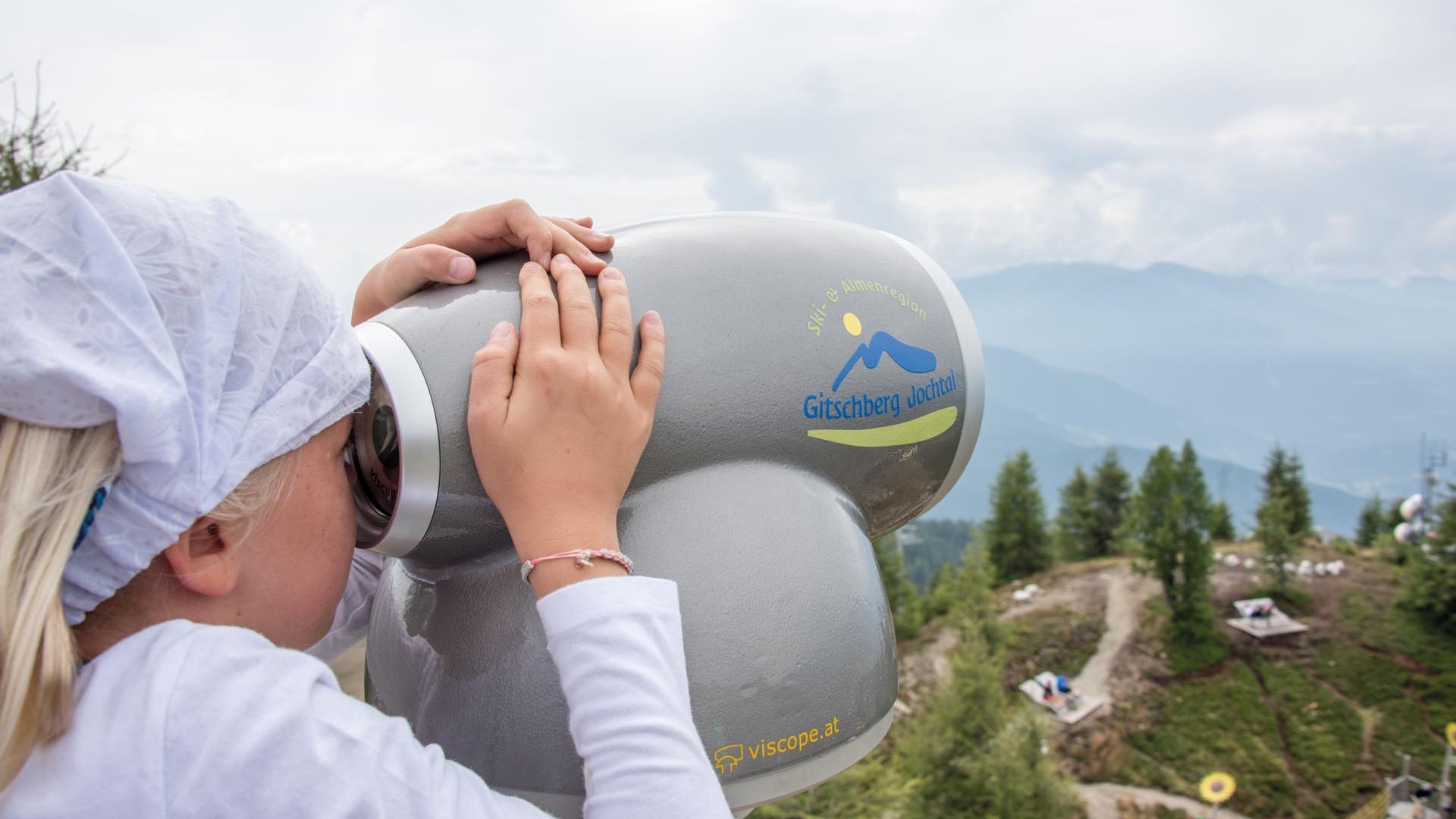 Il Parco del Sole Gitschberg – Maranza L'immagine mostra un bambino con un fazzoletto bianco in testa che guarda attraverso un grande binocolo, posizionato sulla cima di una montagna. Sul binocolo c'è la scritta "Gitschberg Jochtal" e sullo sfondo si vedono montagne nebbiose e un paesaggio escursionistico.
