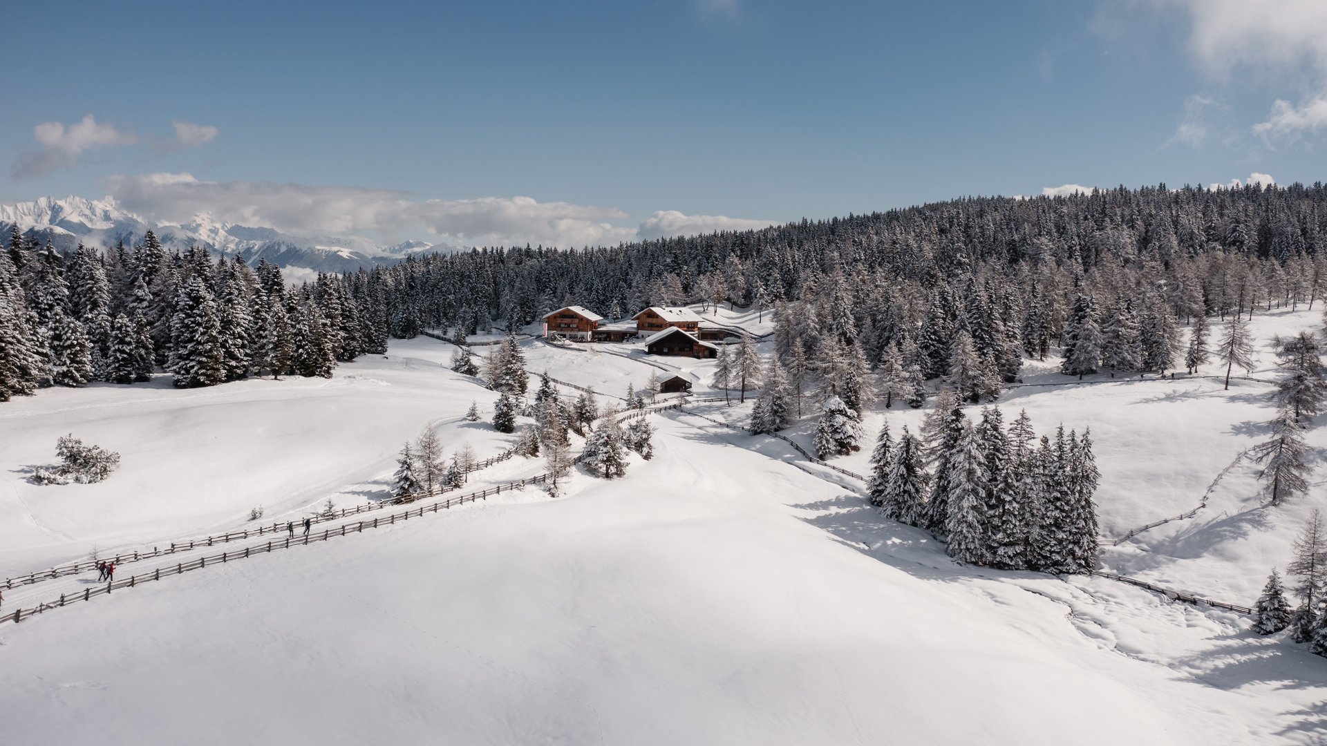 L’Alpe di Rodengo e Luson L'immagine mostra un paesaggio invernale innevato con diverse baite, circondate da uno spesso strato di neve. In primo piano c'è un sentiero innevato che corre lungo una recinzione di legno, e sullo sfondo si estende una fitta foresta di alberi coperti di neve. Il cielo è limpido e azzurro, creando un'atmosfera tranquilla e pacifica.