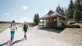 L’Alpe di Rodengo e Luson L'immagine mostra due escursionisti, un uomo e una donna, che camminano lungo un ampio sentiero sabbioso. A destra di loro c'è una piccola cappella di pietra con un tetto in legno. Il cielo è limpido e blu, e sullo sfondo si vedono dolci colline e alberi.