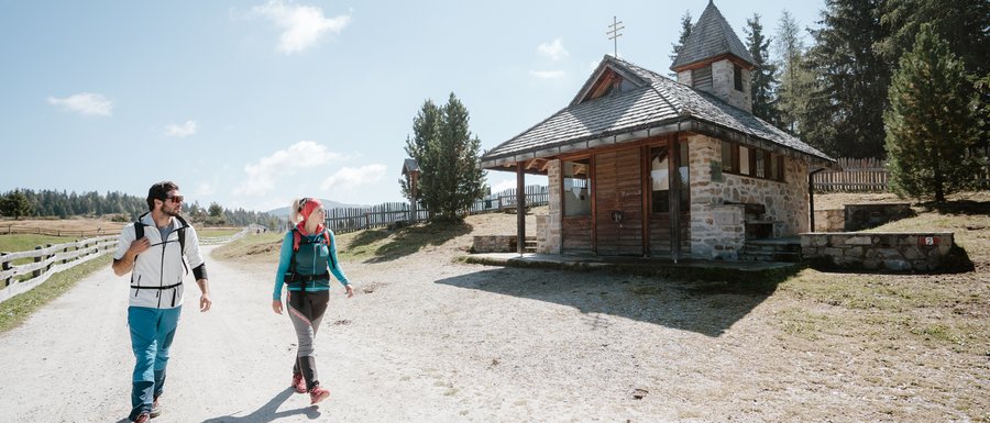 Le località dell’area vacanze Rio Pusteria L'immagine mostra due escursionisti, un uomo e una donna, che camminano lungo un ampio sentiero sabbioso. A destra di loro c'è una piccola cappella di pietra con un tetto in legno. Il cielo è limpido e blu, e sullo sfondo si vedono dolci colline e alberi.